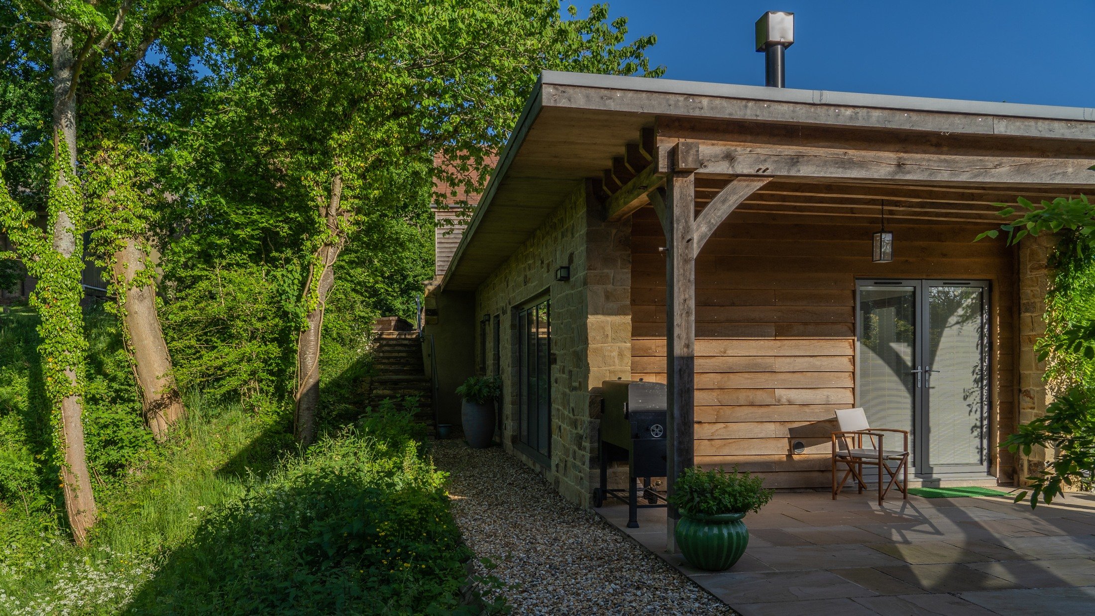 Timber structure, stone walls and a sheltered entrance beside the wooded garden edge.