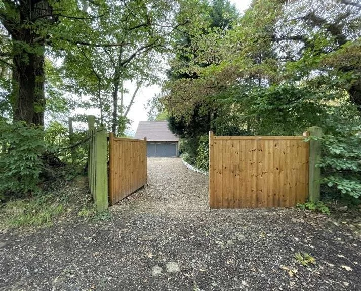 Timber entrance gates open onto a gravel drive leading to the existing garage among mature trees.