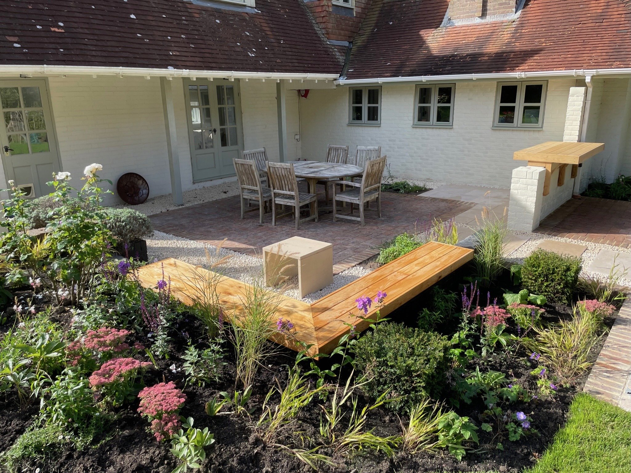 A closer autumn view of timber corner seating among flowering borders, with the dining terrace and timber-topped counter beyond.