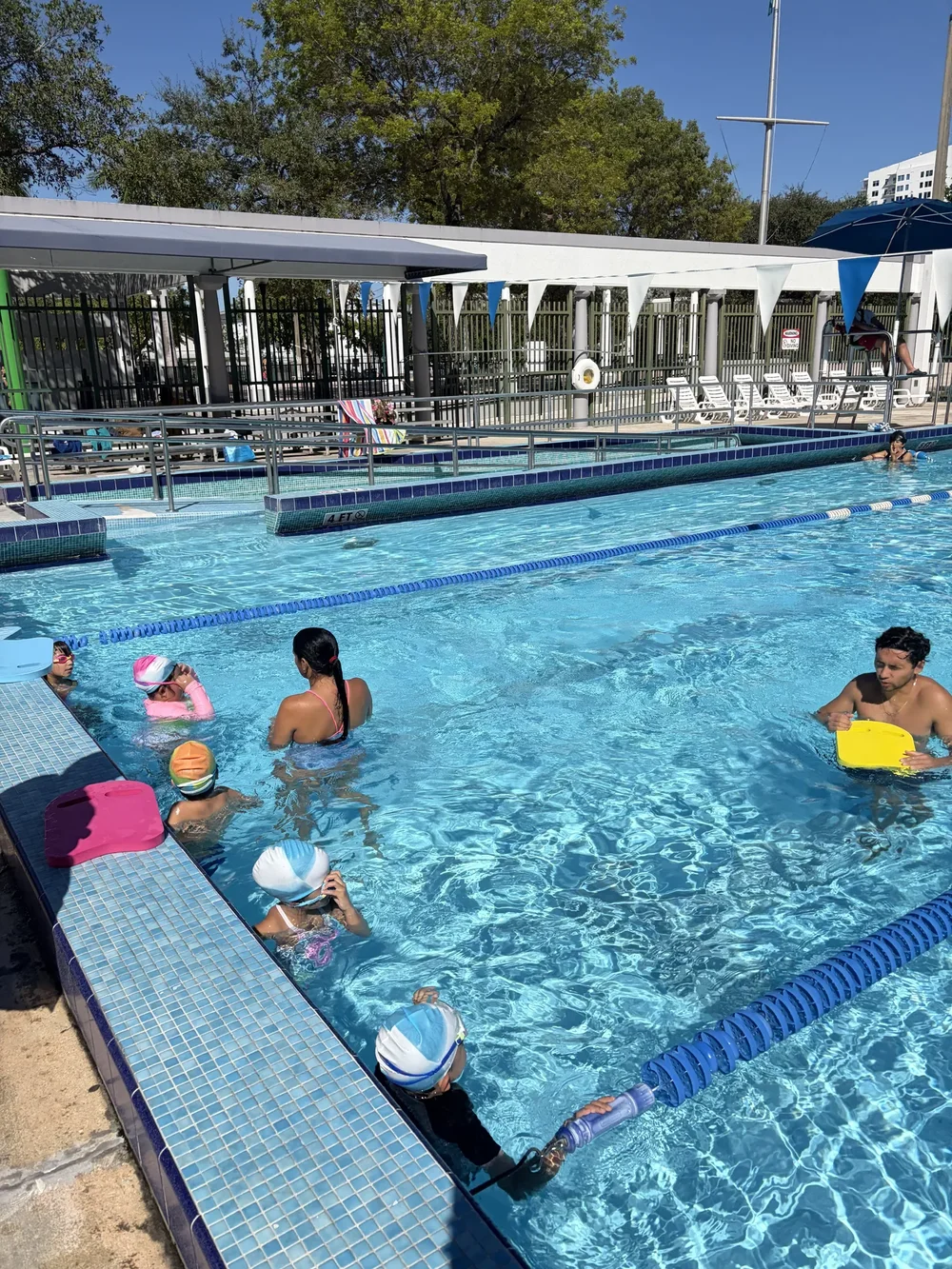 Children wearing swim caps practice with kickboards alongside adults in the pool.