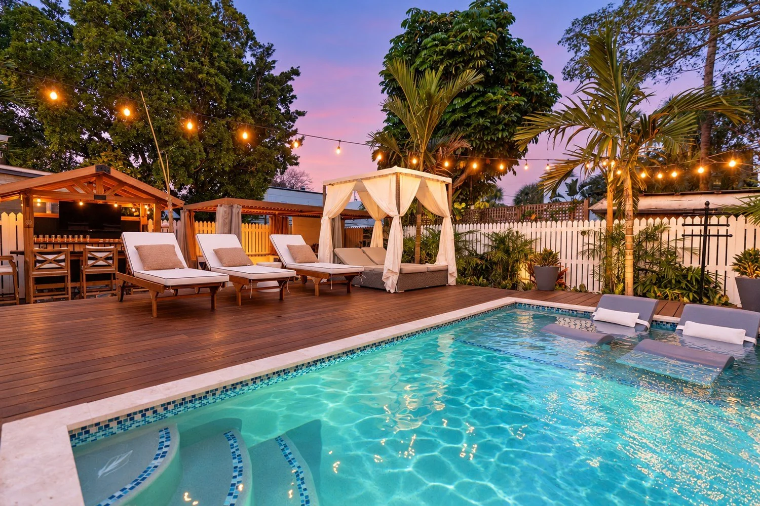 Twilight pool retreat with turquoise water, glowing string lights, a white cabana, and the outdoor bar