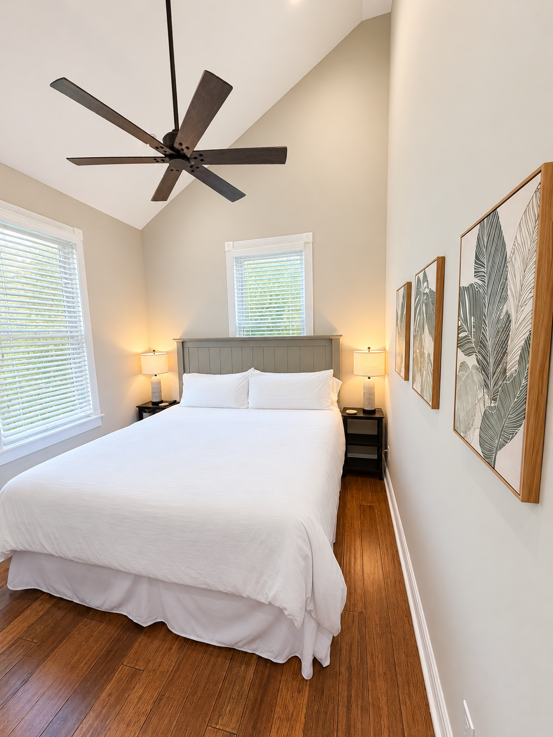 A neatly made bed with white linens and pillows, flanked by nightstands with lamps, in a room with a ceiling fan and artwork on the wall.