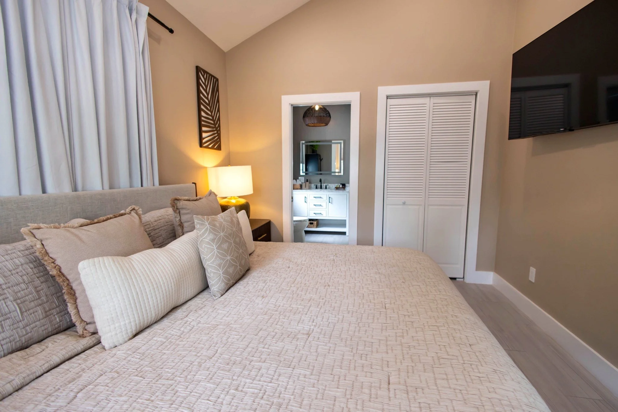 A wide-angle view of a bedroom with a large bed, a TV mounted on the wall, and a lamp on a nightstand.