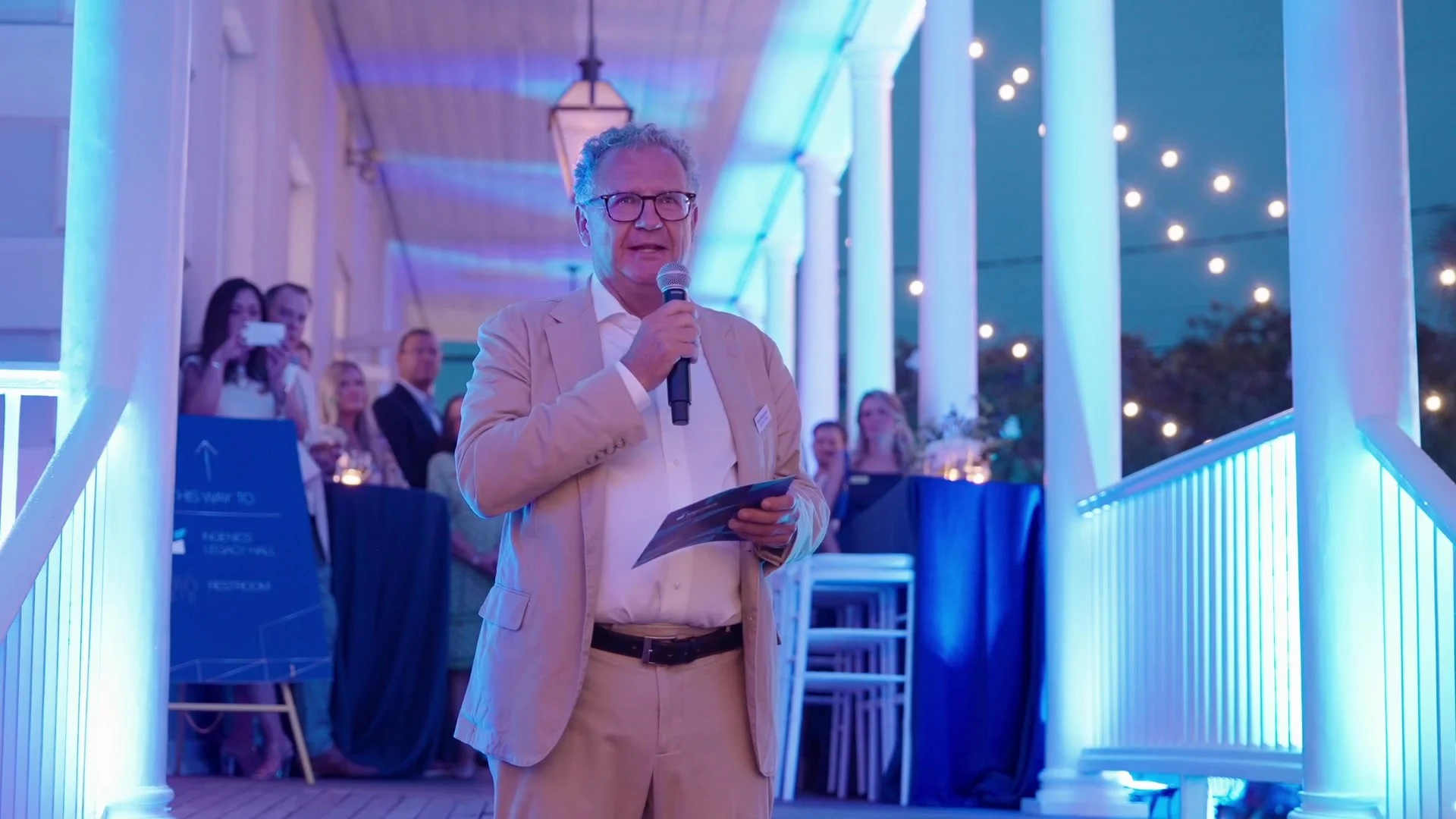 A speaker at Ingenics’ anniversary, framed against blue-lit columns and warm evening lights