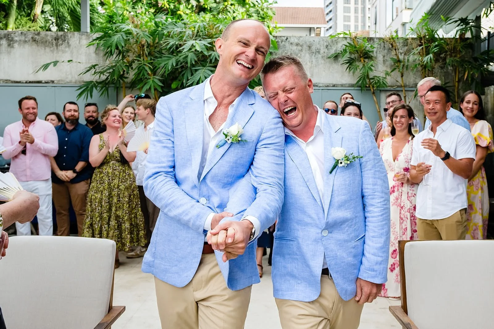 Two grooms in pale blue jackets laughing together, surrounded by their guests.