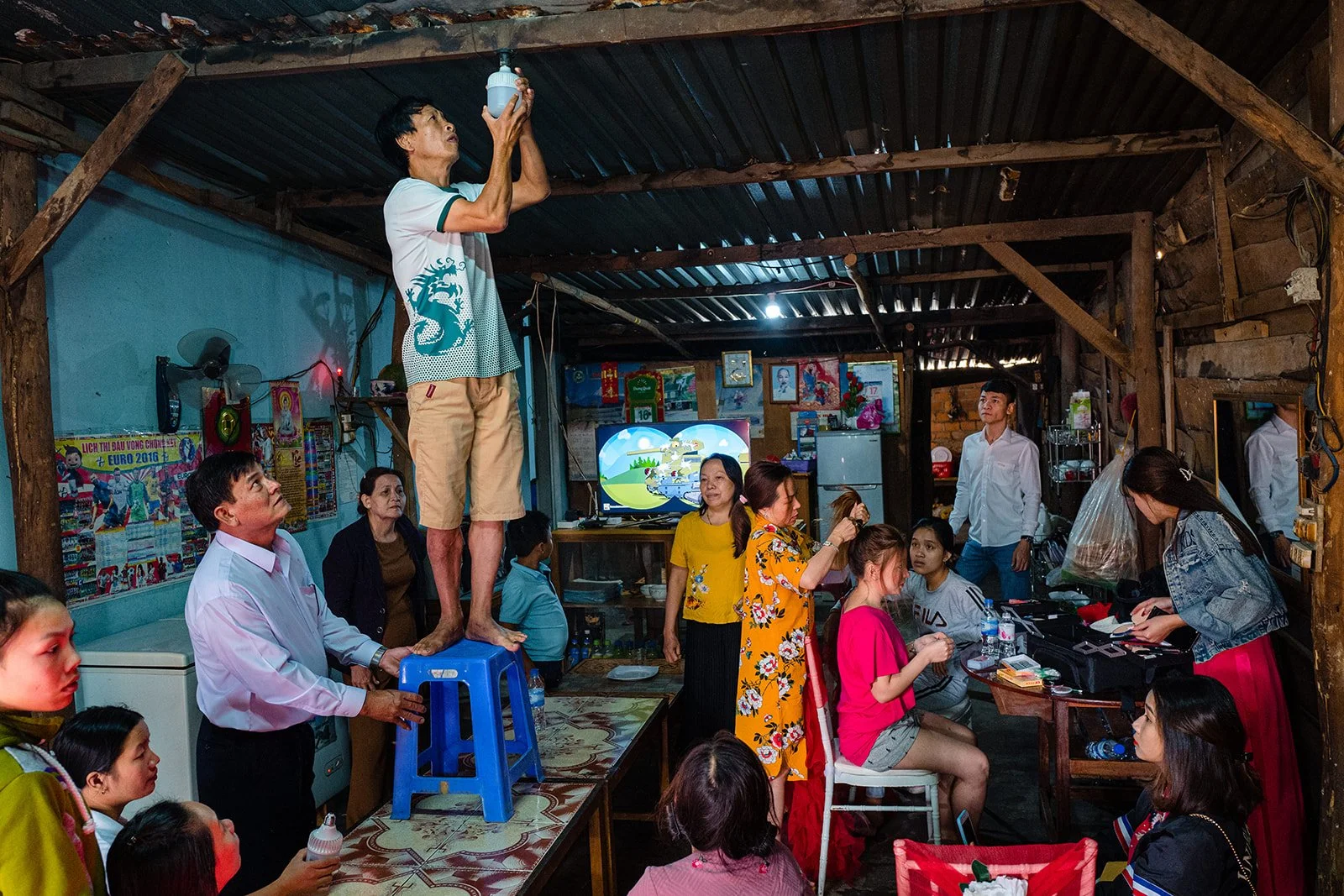 A busy Vietnamese home on a wedding morning, with family preparing and a man reaching up to change a light bulb.
