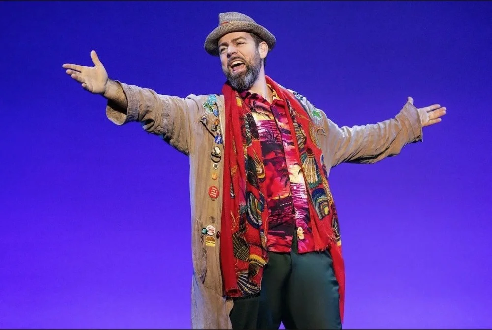 The Happy Man, in a patched coat and fedora, sings with arms spread wide in the Pretty Woman tour