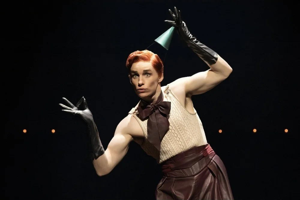 Eddie Redmayne as the Emcee in a party hat and long black gloves