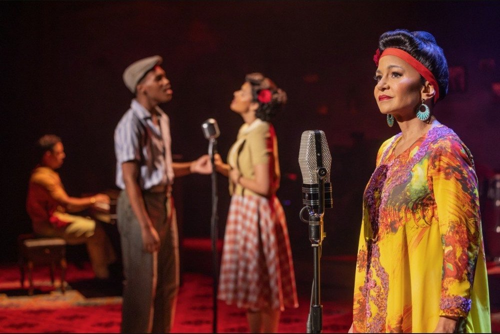 A singer in a yellow dress at a vintage microphone, with performers singing behind her, in the Buena Vista Social Club North American tour