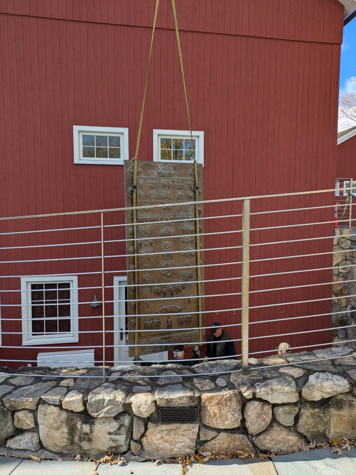 A bronze panel being lowered on straps beside the red Visitor Center, over the stone wall and cable railing of the stairwell.