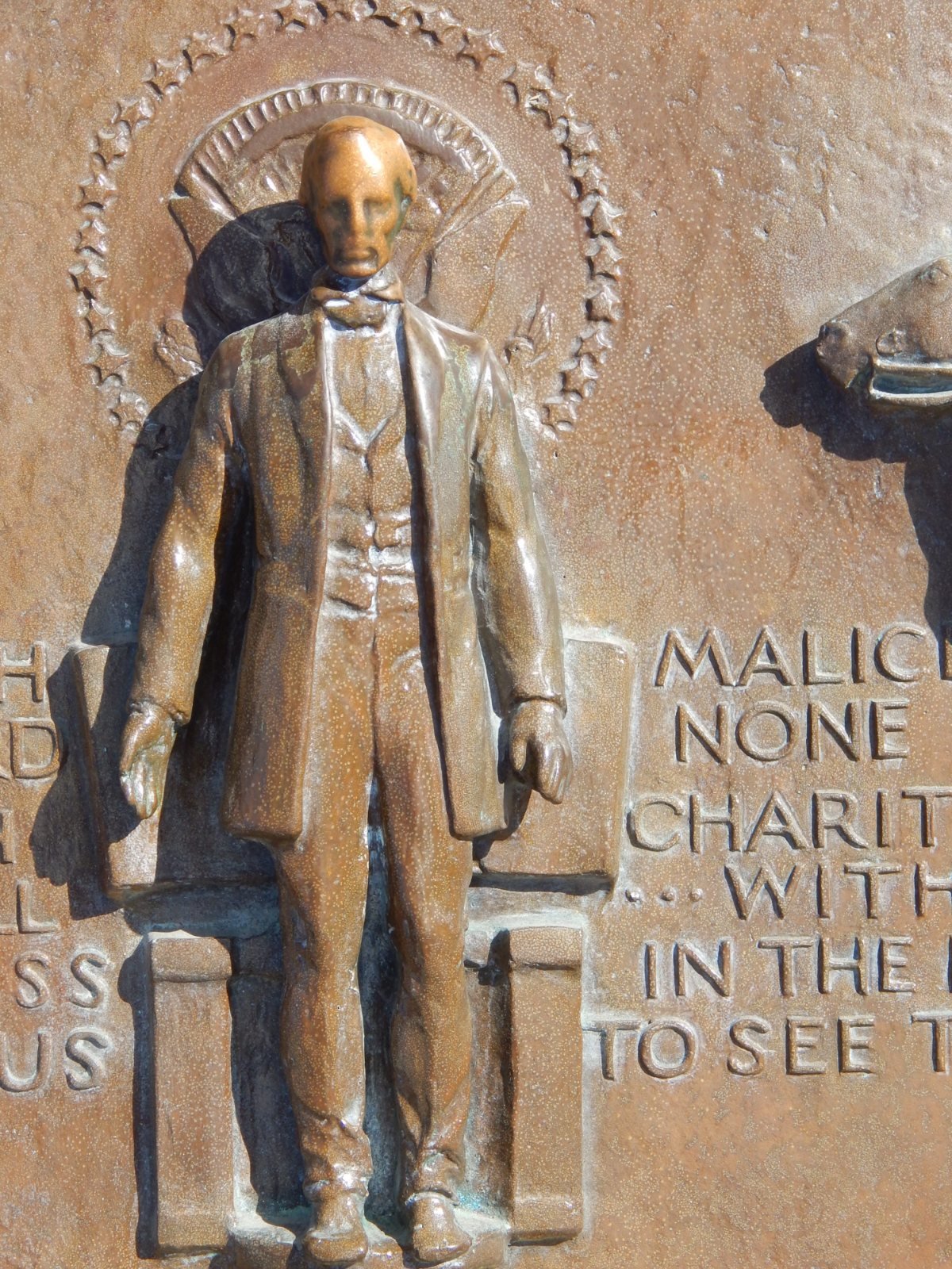 Close-up of the bronze figure of Abraham Lincoln standing before a ring of stars, his face polished bright against the darker bronze, beside the carved words With malice toward none, with charity for all.