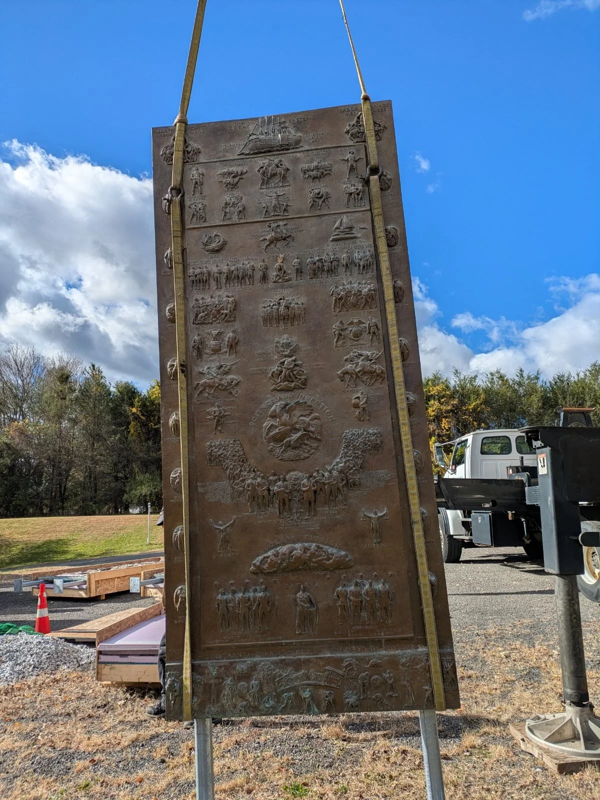 One bronze panel hanging upright from a crane's straps against a blue sky, with the crane truck behind.