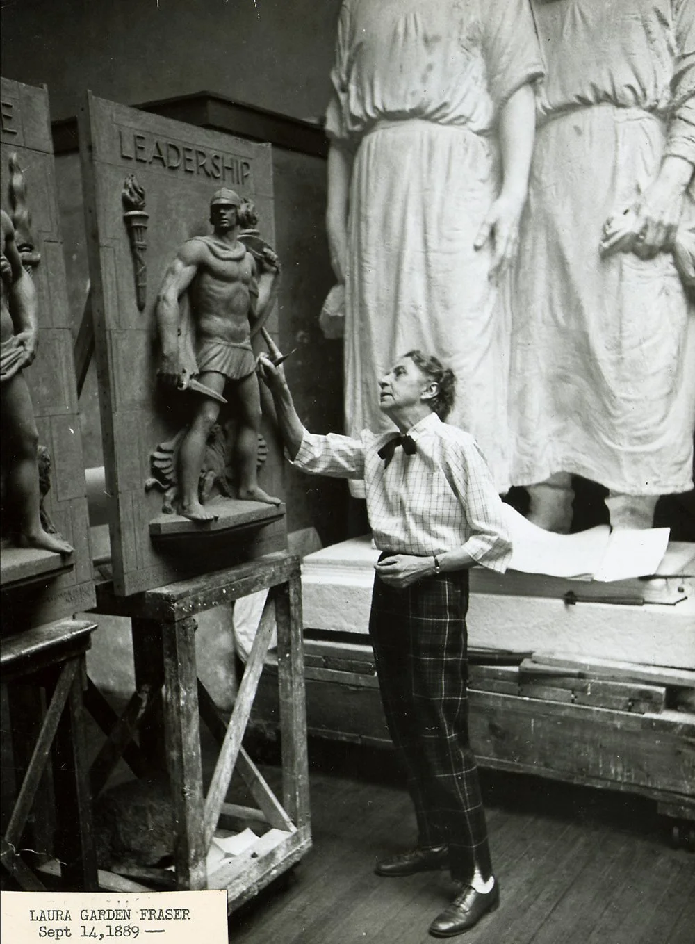 Laura Gardin Fraser, white-haired, in a checked shirt and bow tie, reaching up to a tall relief of a warrior titled Leadership in her studio, with large plaster figures behind her.