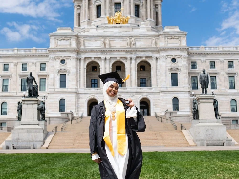 A graduate in a black gown and gold stole in front of the Minnesota State Capitol