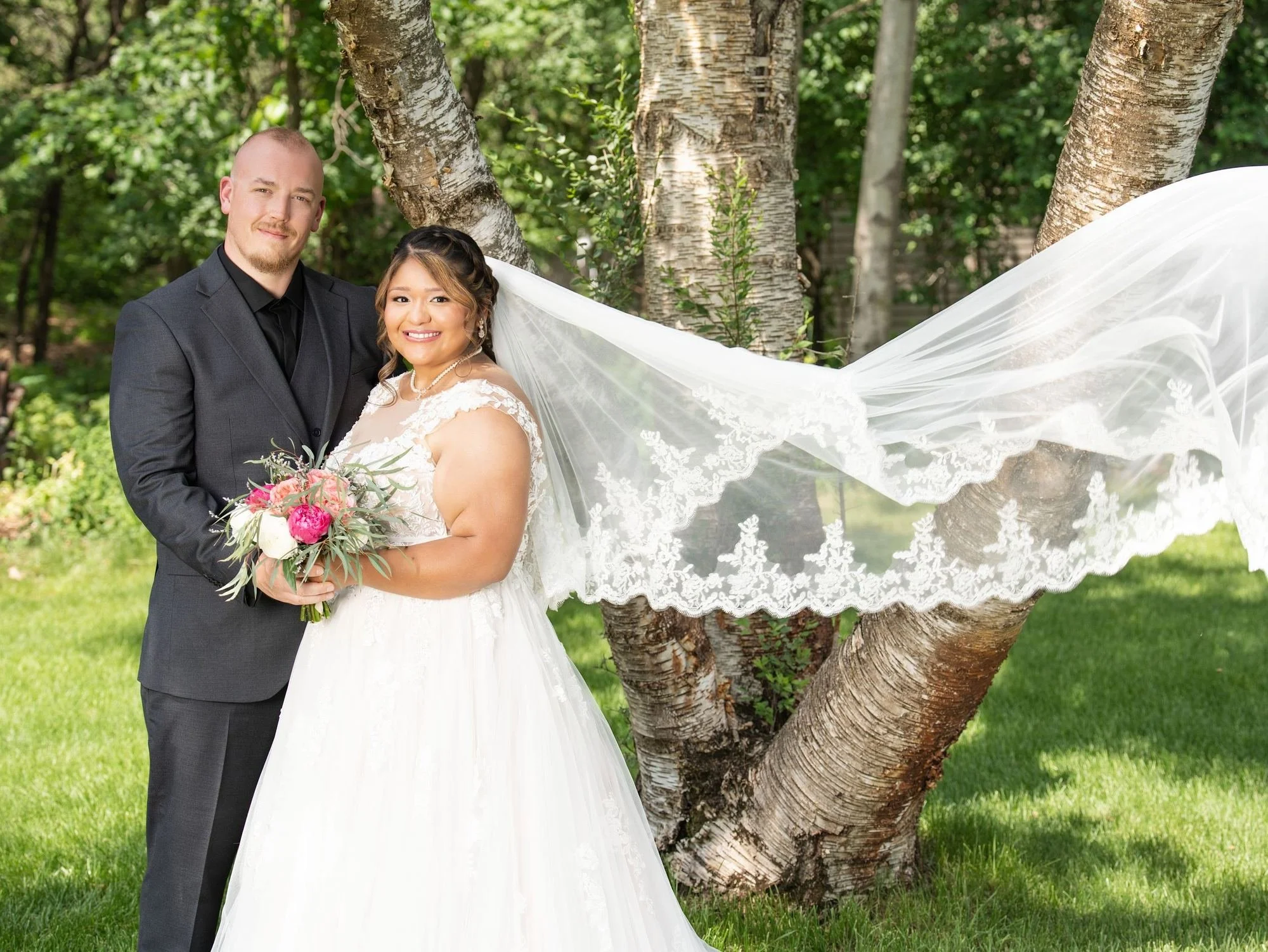 A bride's long lace veil billows in the wind as she stands with the groom under birch trees