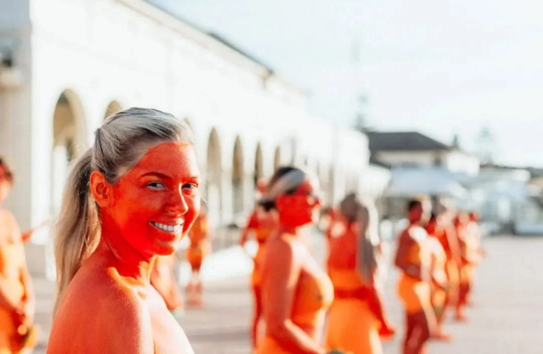 A smiling woman with orange-painted skin looks over her shoulder as a line of people stand along a seaside promenade