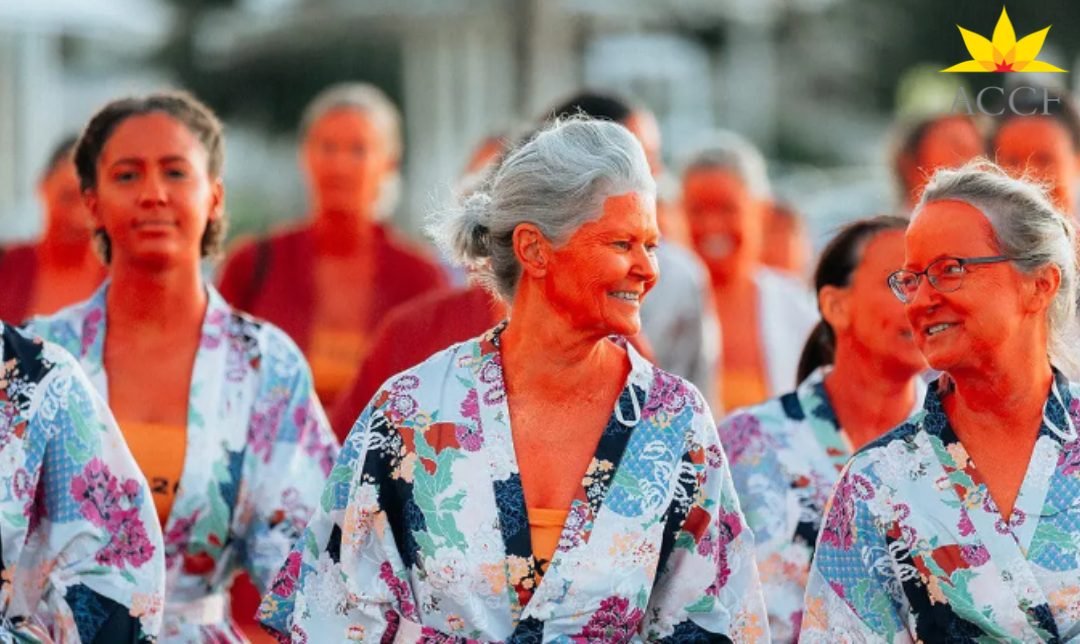 Women of different ages in floral kimono-style robes smile and walk together, their skin painted orange