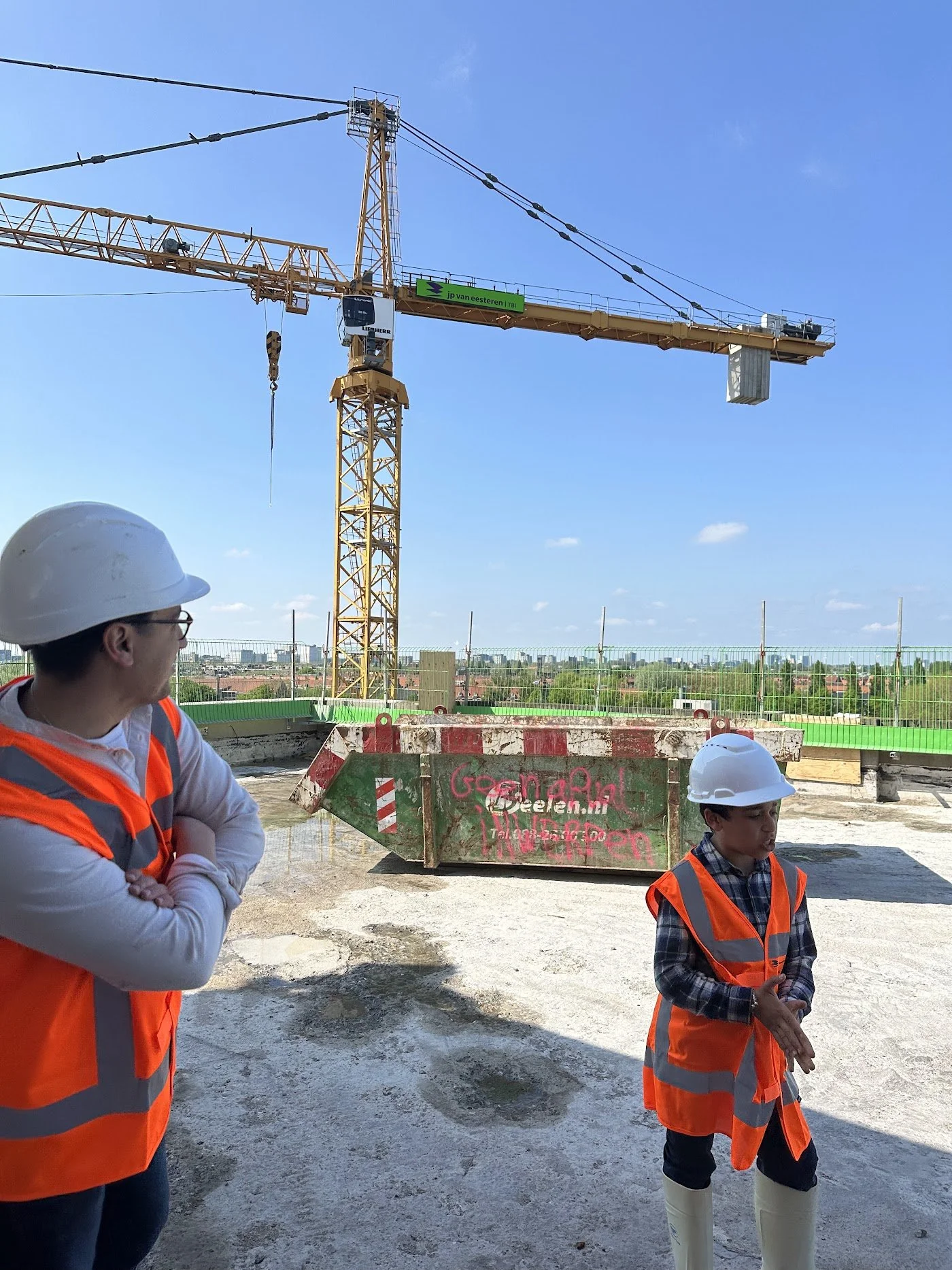Driss in a hard hat and safety vest on a construction site, looking up at a crane