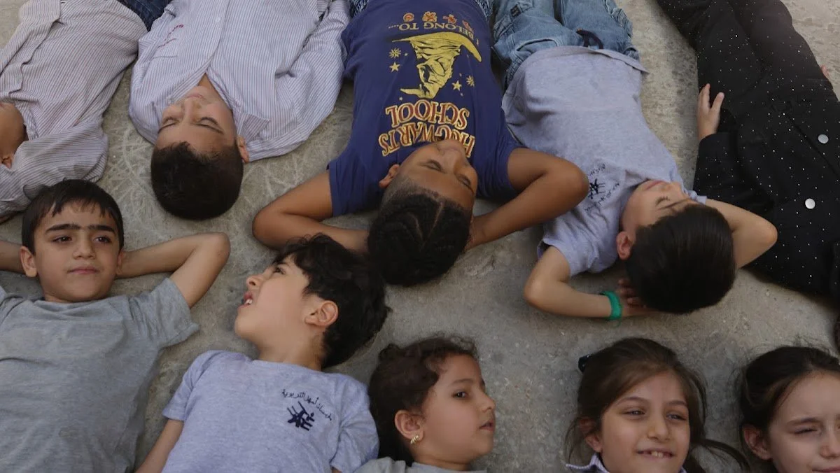 Children lying in a circle on the schoolyard, smiling up at the camera