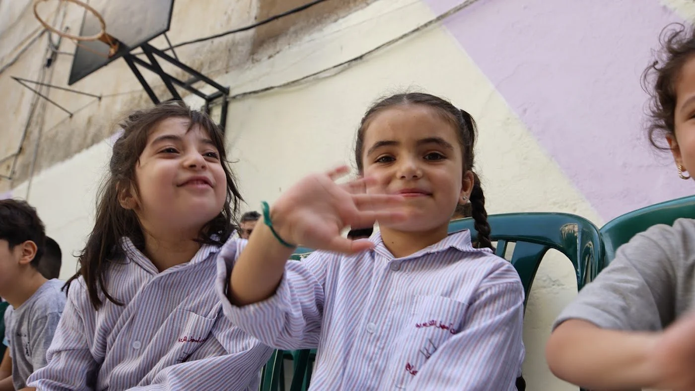 Two girls in school uniforms waving and laughing during a Make A Kid Happy visit in Lebanon