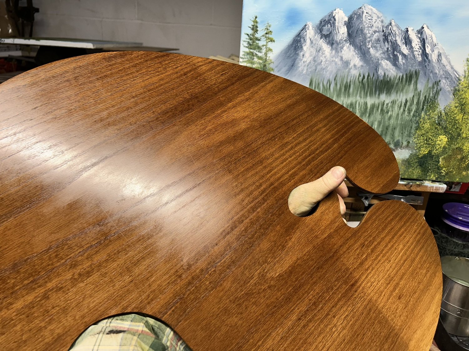 One of Johnny Harris's handmade wooden palettes with a glossy finish, held in front of a mountain painting