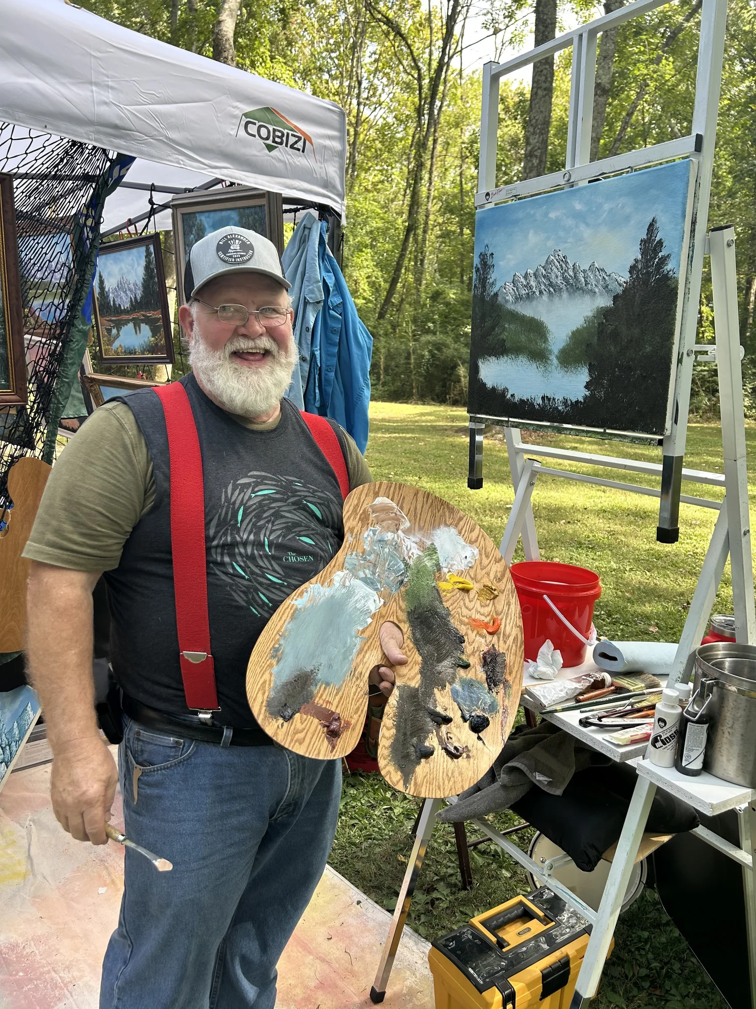 Johnny Harris smiling with his paint palette beside an easel and a mountain landscape painting