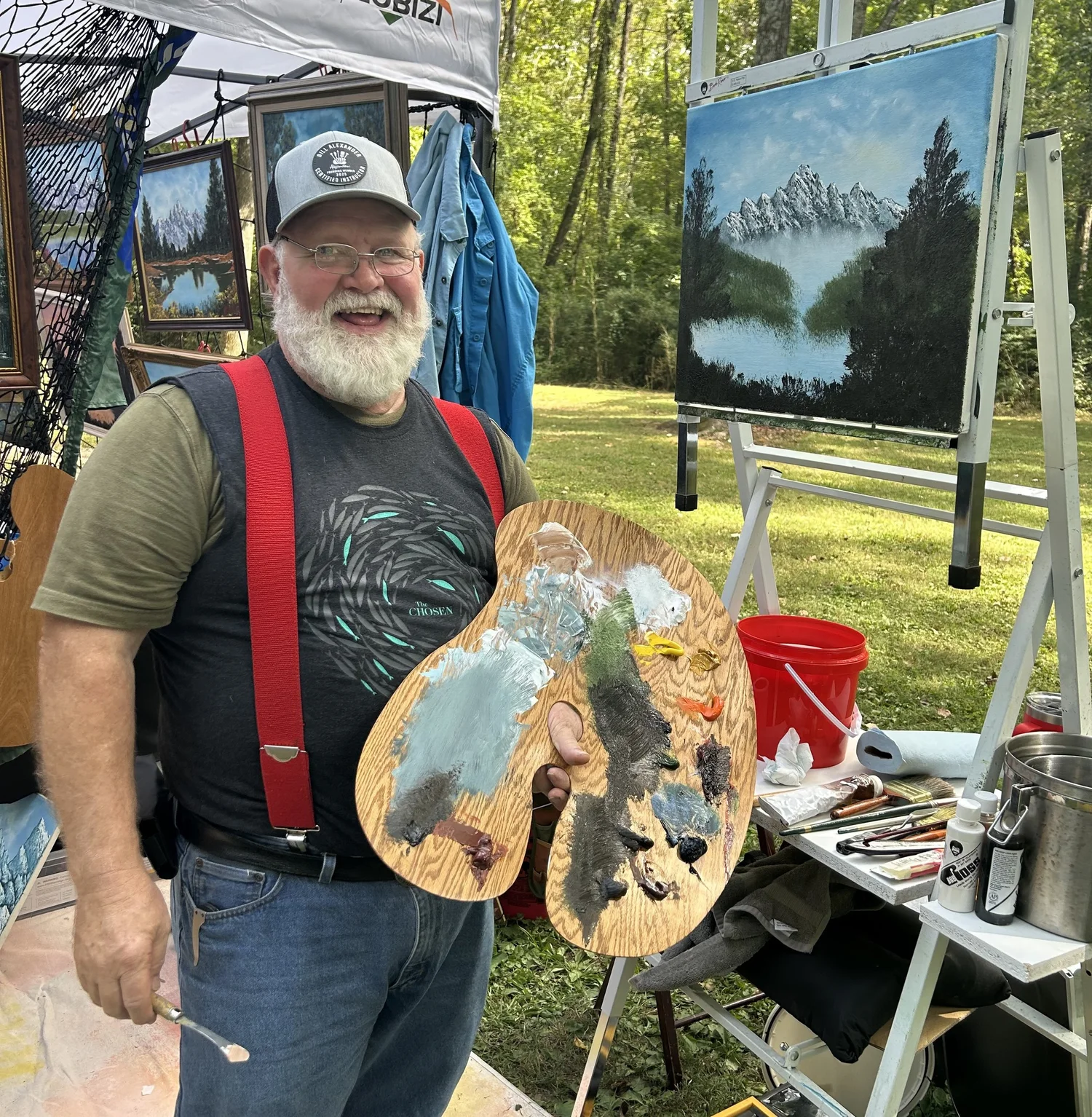 Johnny Harris smiling with his palette beside a mountain painting at his art fair booth