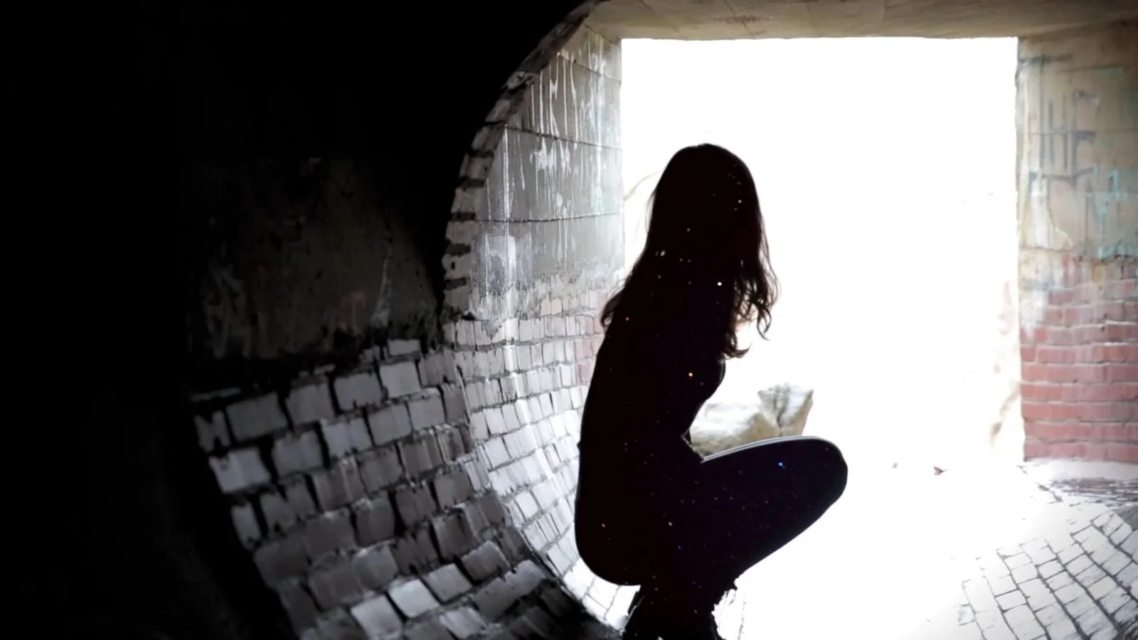 A woman in silhouette crouches at the bright end of a brick tunnel