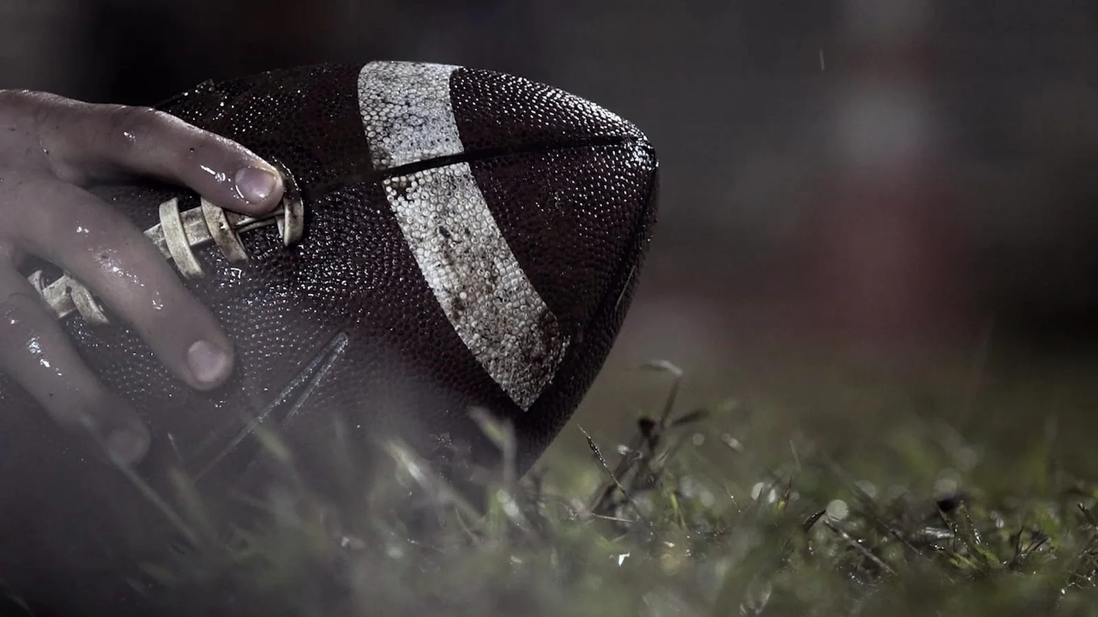 A hand grips an American football in wet grass