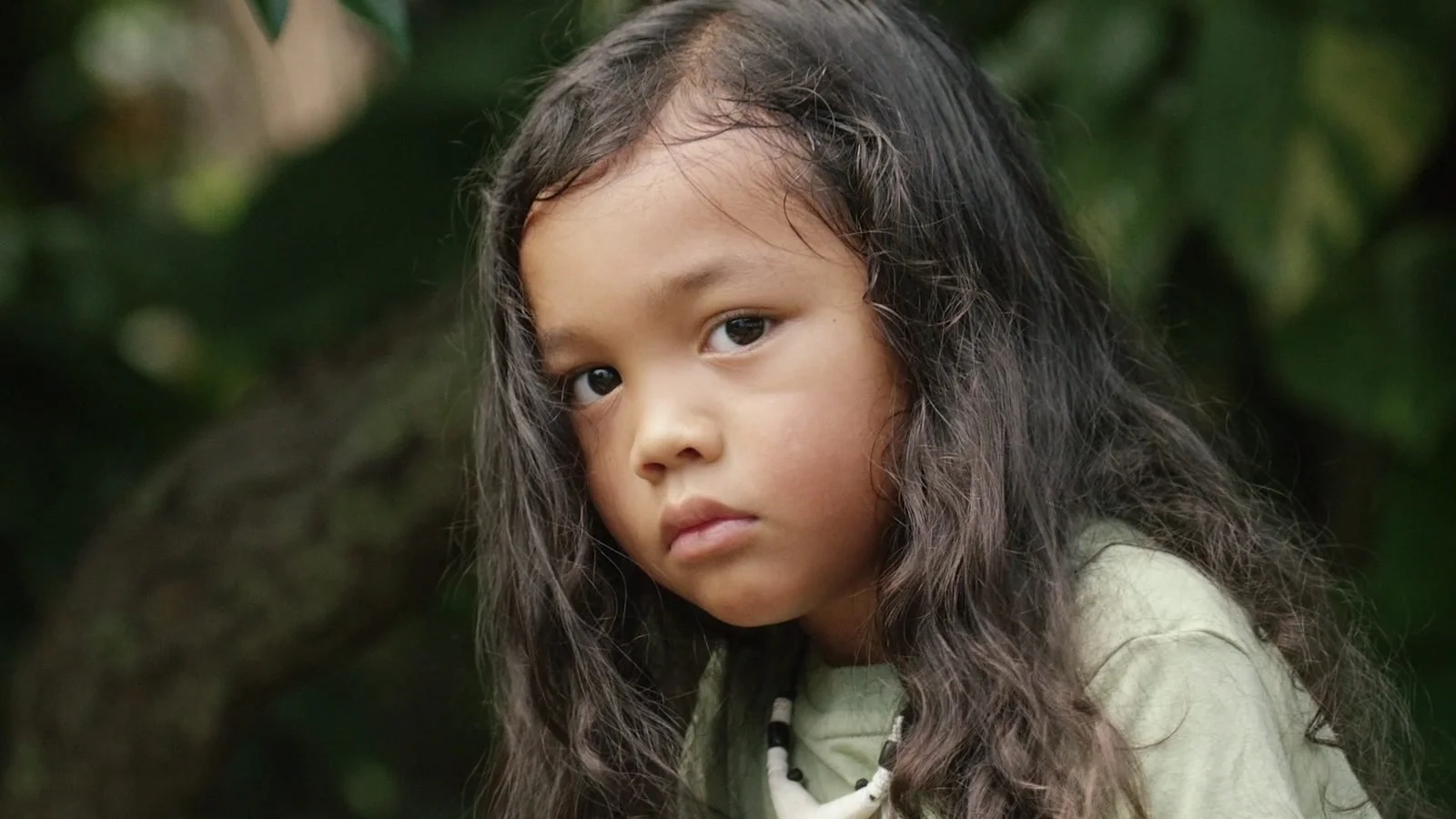 A young girl with long dark hair looks into the camera in a Guam forest