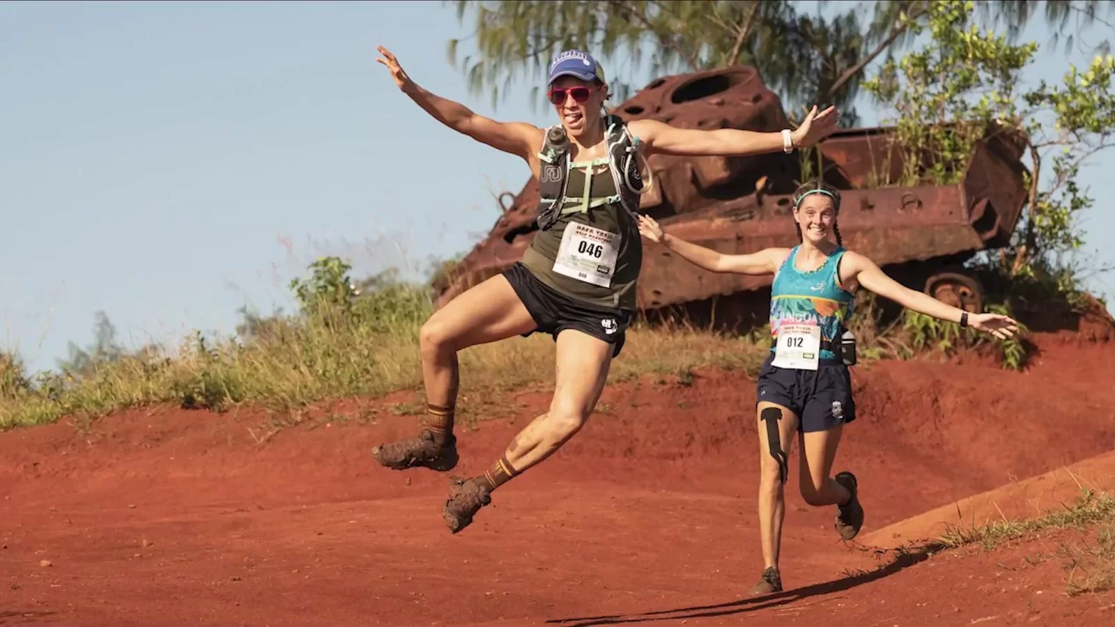 Two trail runners leap for joy on a red dirt path in front of a rusted tank