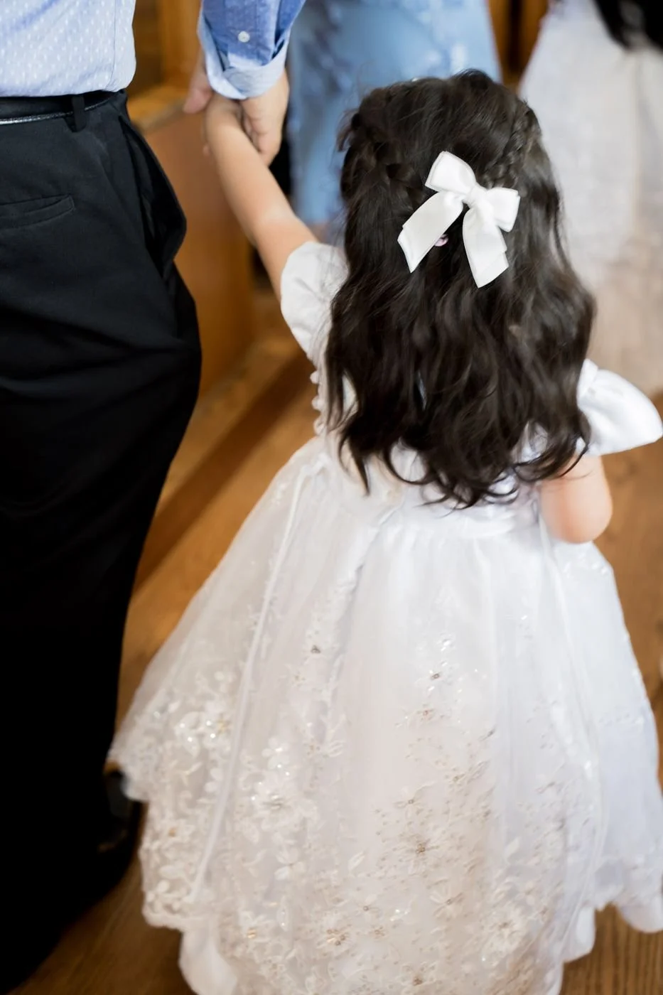A little girl in a white baptism gown with a satin bow, seen from behind