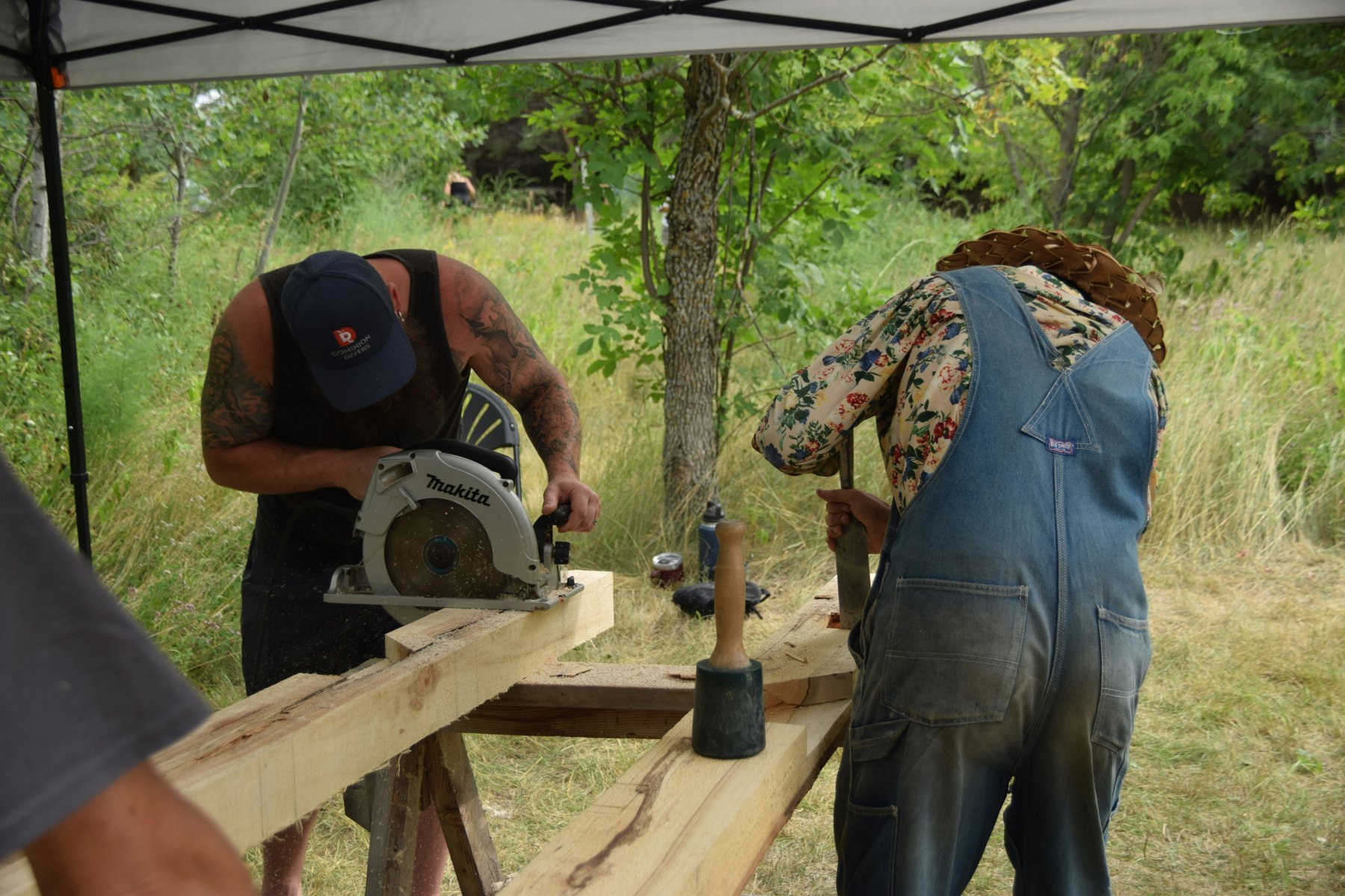 Zwei Handwerker bearbeiten gemeinsam Holzbalken mit Kreissäge und Stemmeisen im Freien