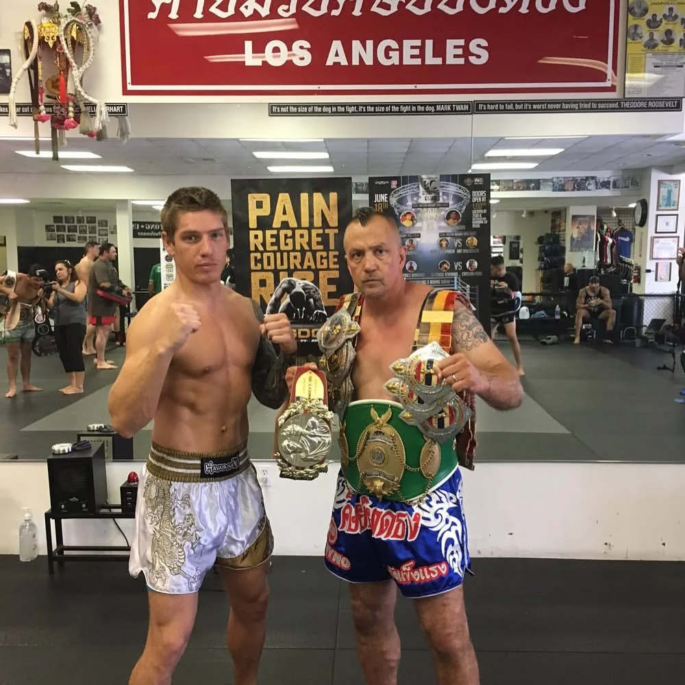 Daniel posing with a Muay Thai coach wearing championship belts