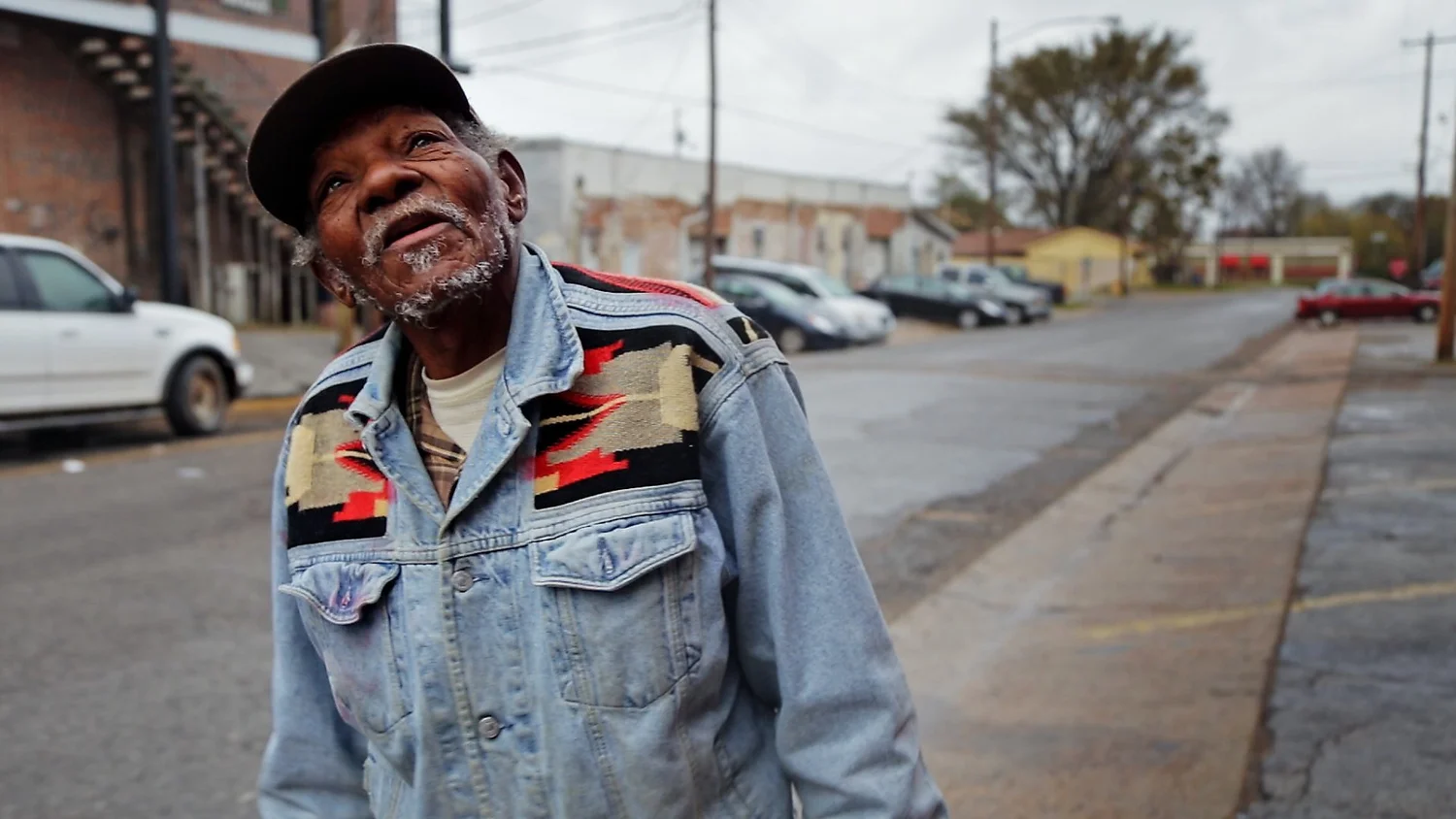 An older man in a cap and a denim jacket with a red, black and cream patterned yoke smiles and looks up, standing on a quiet street lined with parked cars.