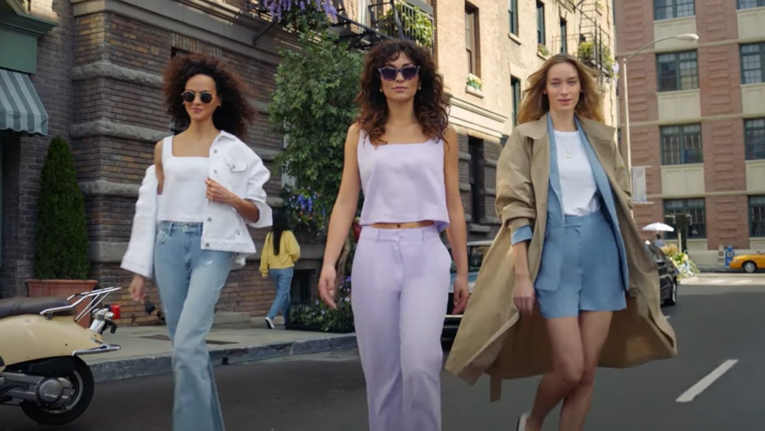 Three women walk side by side down a sunny city street of brick buildings: One in a white denim jacket and light jeans, one in a lavender crop top and trousers with purple sunglasses, and one in a light blue shirt and shorts under a long tan trench coat.
