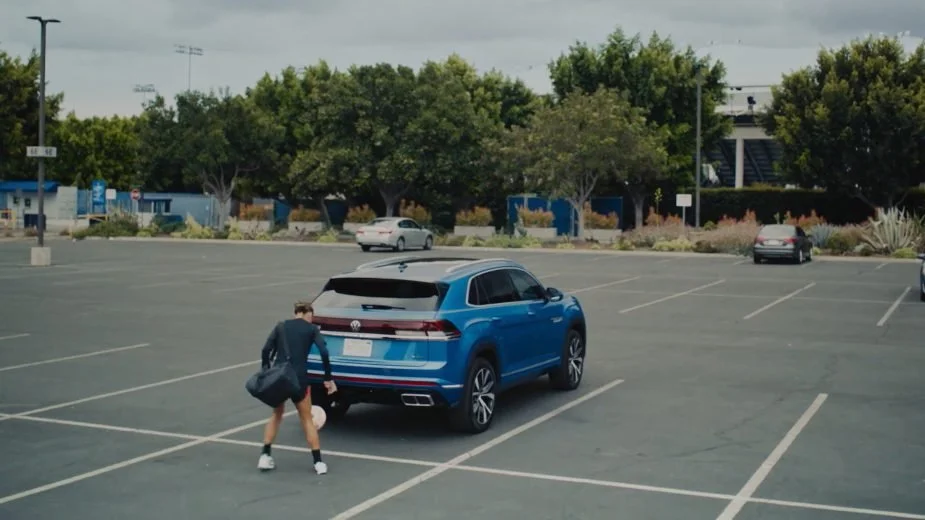 Seen from behind, a soccer player in a dark top and shorts, a duffel bag on her shoulder, keeps a ball in the air beside a blue Volkswagen SUV in a nearly empty parking lot lined with trees.