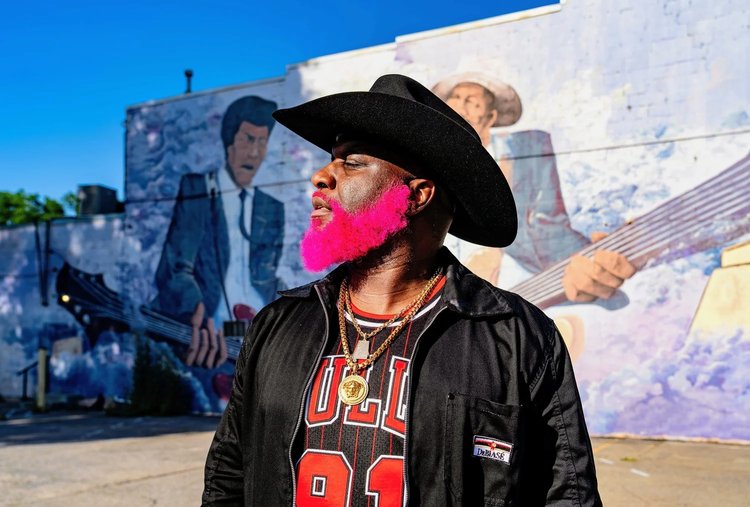 Pynk Beard, with a bright pink beard, a black cowboy hat, gold chains and a black jacket over a red and black jersey, looks off to one side in front of a sunlit mural of guitar players.