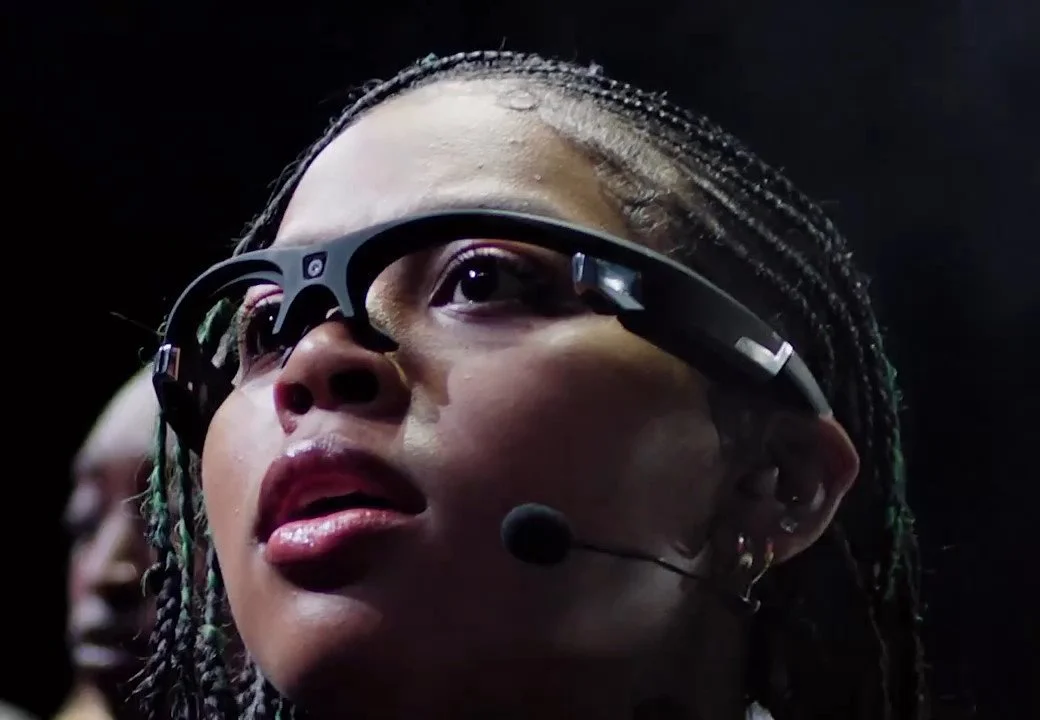 Close-up of a woman with long braids wearing futuristic sensor glasses and a headset microphone, lit against black.