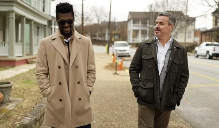 Two men walk side by side along a residential sidewalk on an overcast day: One in sunglasses and a long camel coat, smiling, and one in a dark field jacket over a white shirt and gray sweater.