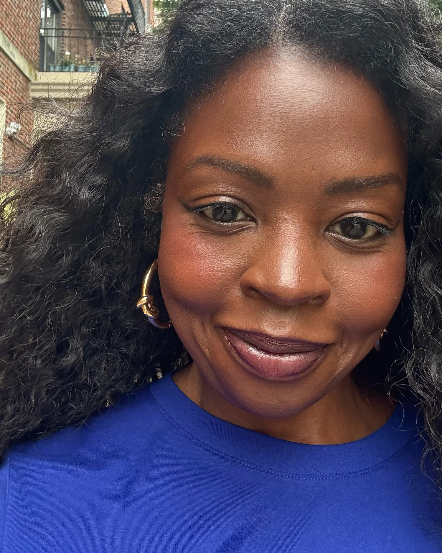 Jocelyn Michelle Brown smiling at the camera in a cobalt-blue top and gold hoop earrings, her dark hair full around her face, on a city street with trees and brick buildings behind her.