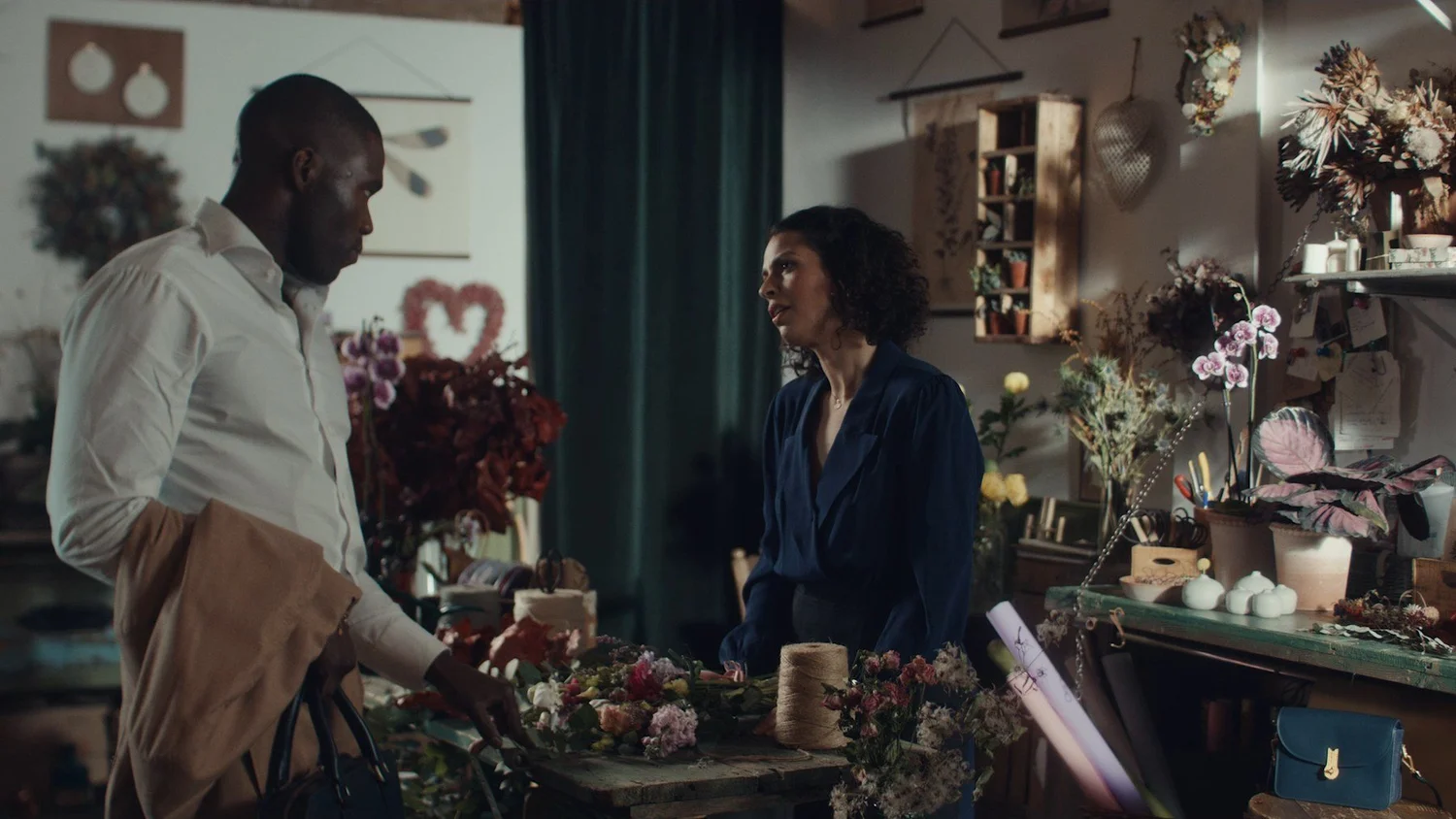 In a flower shop, a man in a white shirt holds a jacket over his arm as a woman in a navy blouse looks over at him across a worktable covered in flowers.