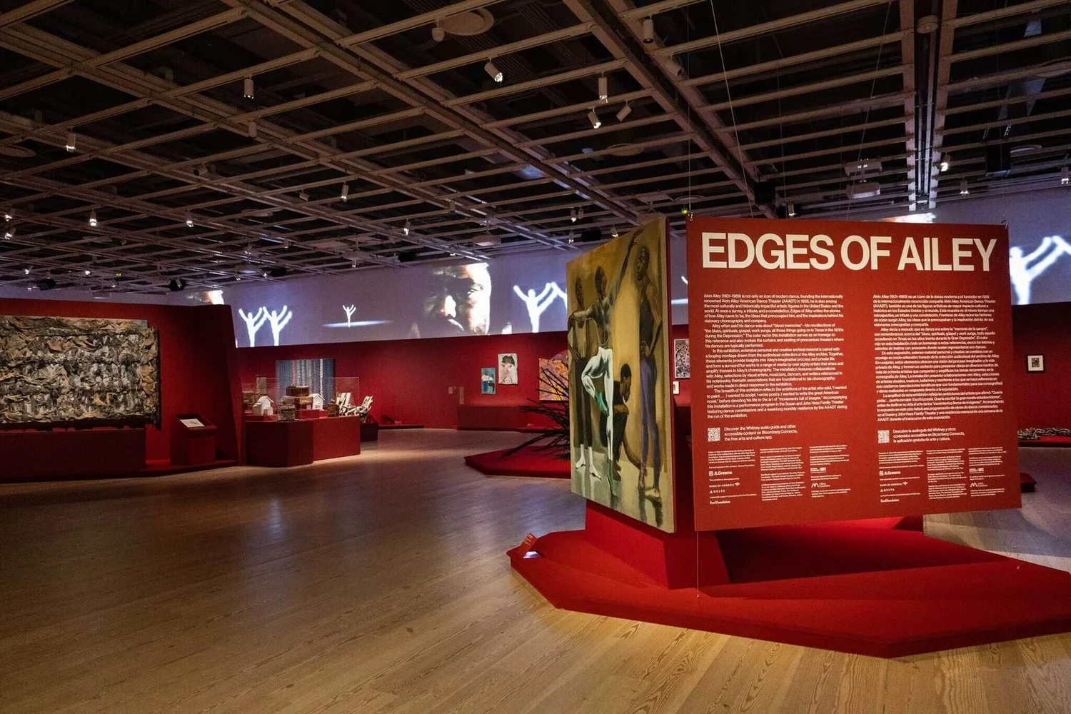Installation view of Edges of Ailey at the Whitney: A red title wall and a painted panel stand in the foreground, while large projected images of Alvin Ailey and dancers run along the gallery’s upper walls.