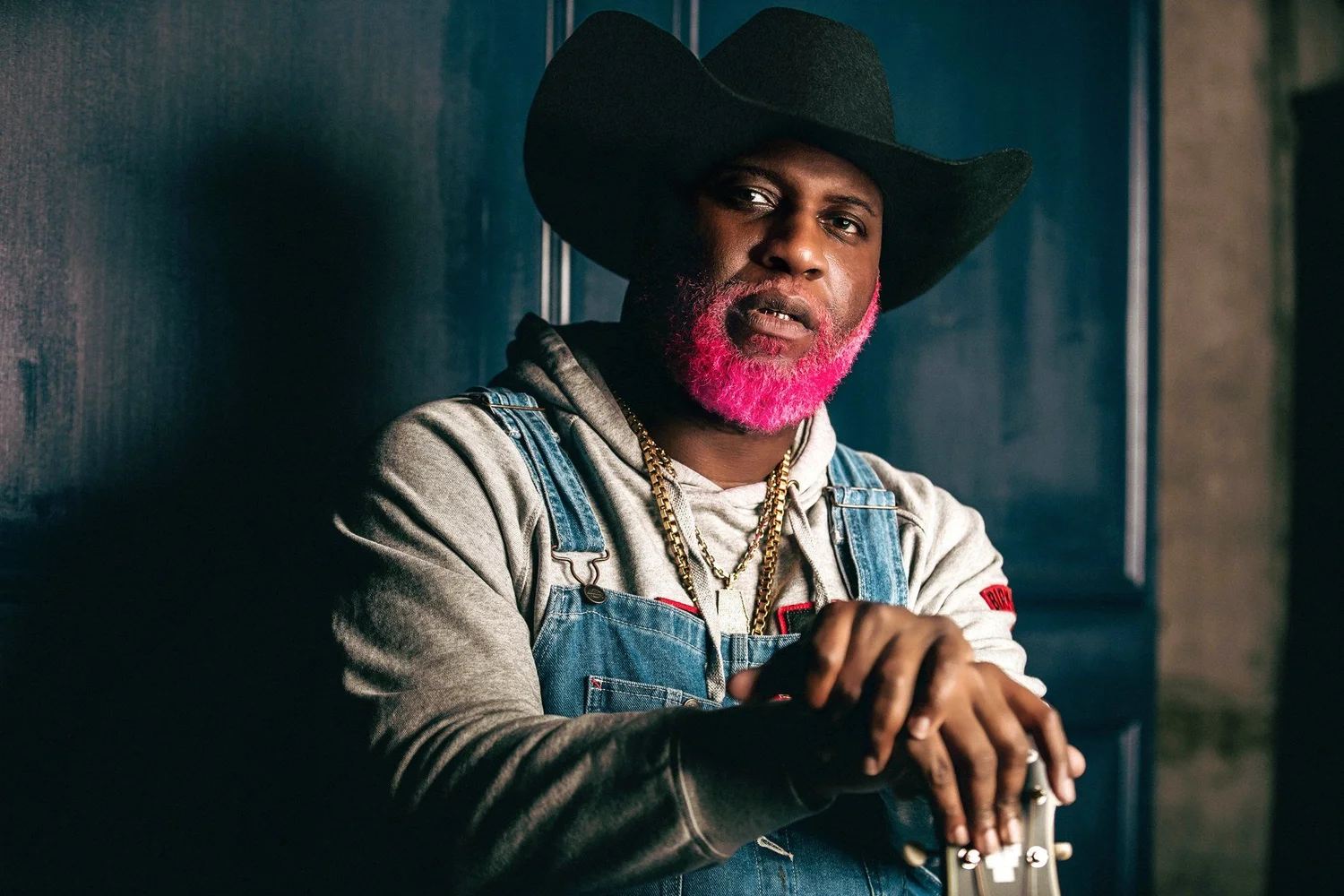Pynk Beard, with a bright pink beard and a black cowboy hat, wears a grey hoodie under denim overalls and gold chains, leaning against a dark blue wall with his hands resting on the neck of a guitar.