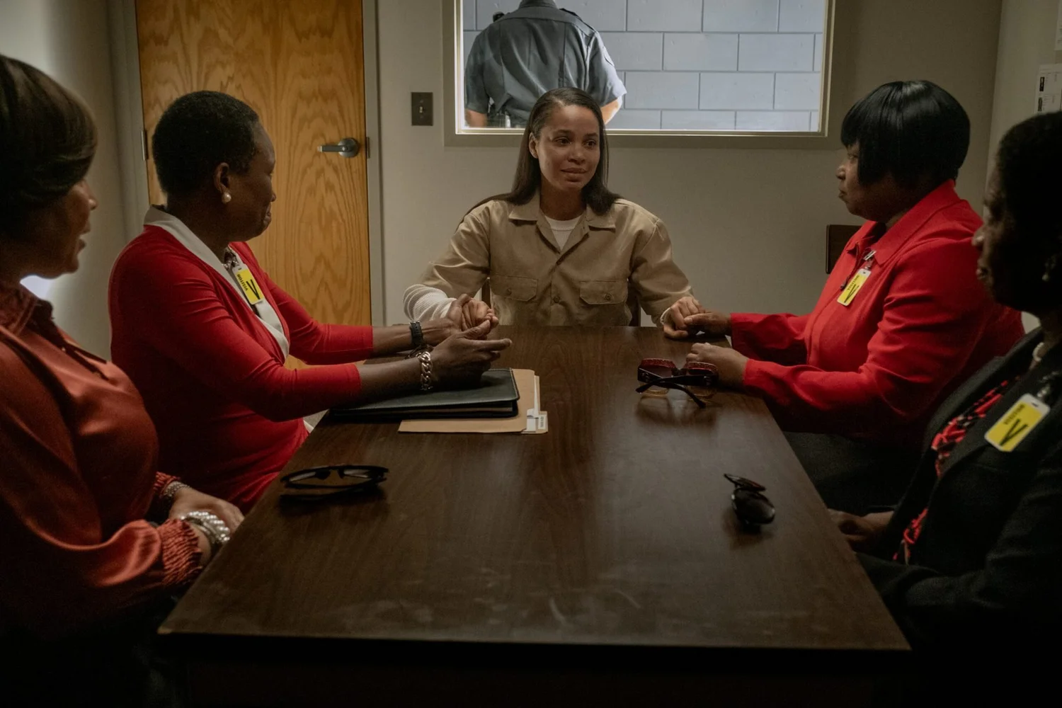 A young woman in a tan prison uniform sits at the head of a wooden table, holding hands with women in red jackets and visitor badges seated on either side, while a guard stands at the window behind her.