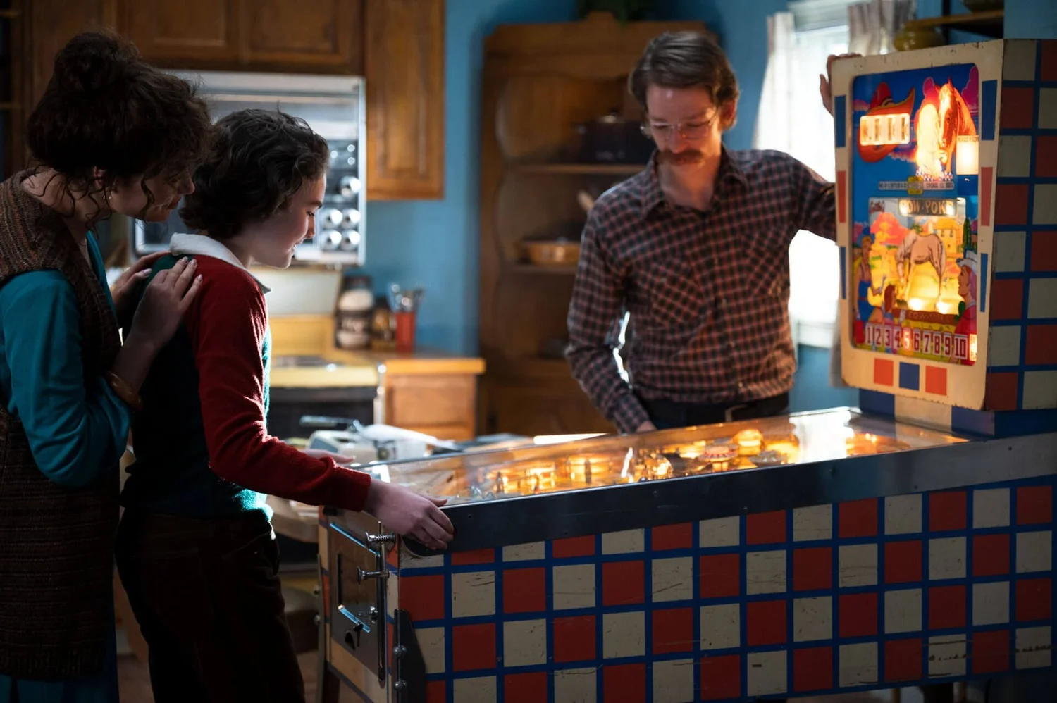A family gathers around a pinball machine in a 1970s kitchen: A boy works the flippers while a woman rests her hands on his shoulder, and a mustached man in glasses and a plaid shirt leans over the far side of the playfield.