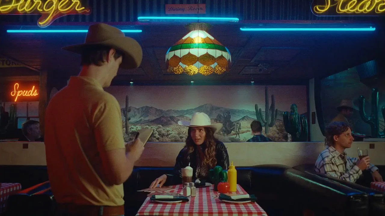 A diner scene from Ally’s Life Today campaign: A waiter in a cowboy hat takes an order from a woman in a white cowboy hat at a booth with a red gingham tablecloth, under a stained-glass lamp and neon signs.