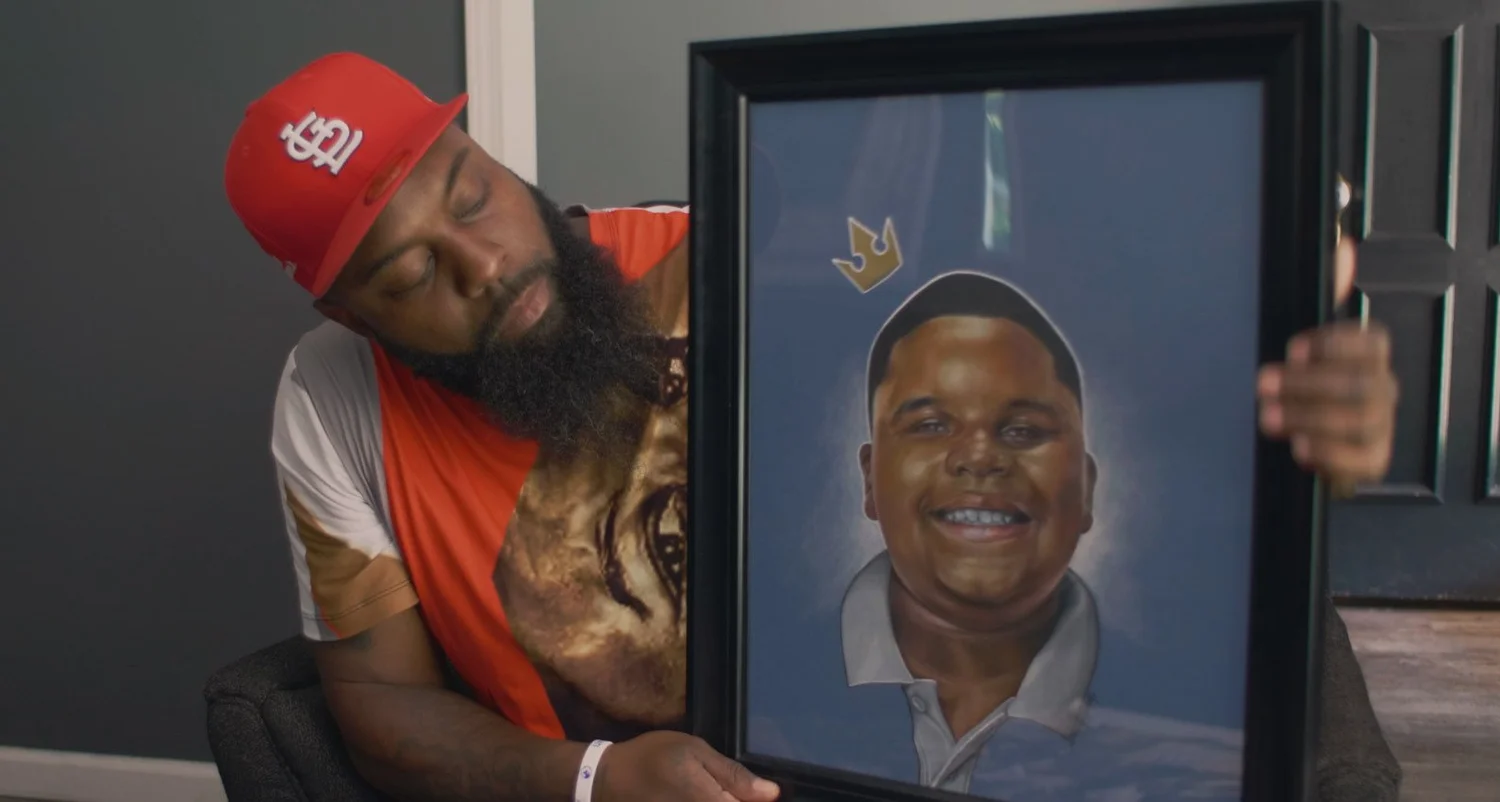 Michael Brown Sr., in a red St. Louis Cardinals cap, looks down at a framed painted portrait of his son, Michael Brown Jr., who is smiling beneath a small gold crown.