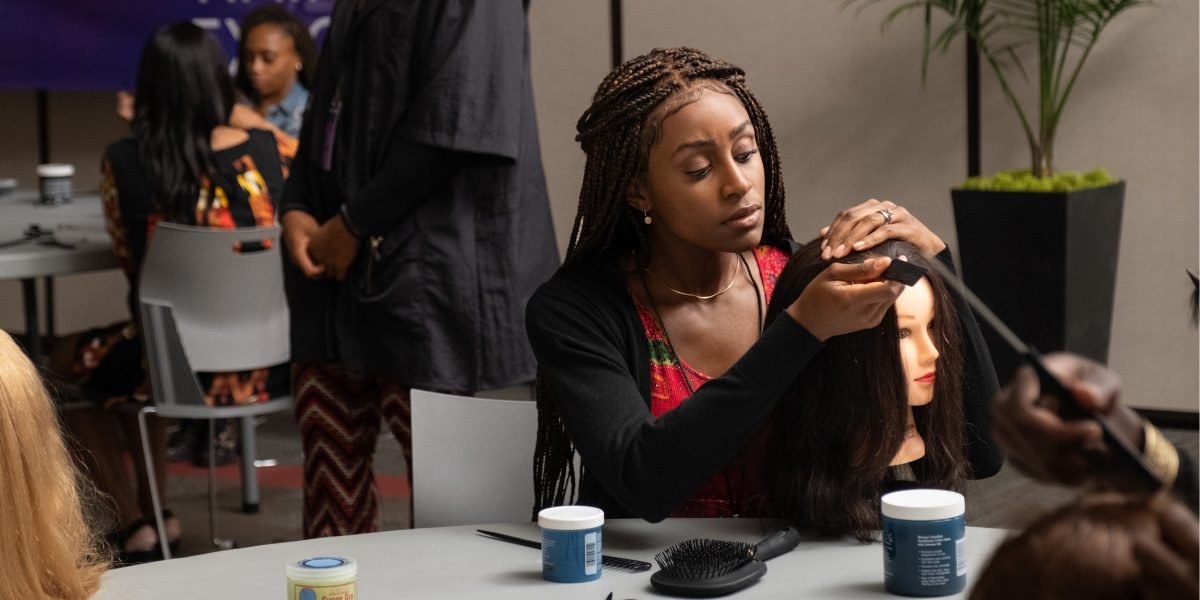 A young woman with long braids, in a black cardigan over a red patterned top, parts the hair of a mannequin head at a table set with a brush, a comb and jars of hair product, in a busy classroom.