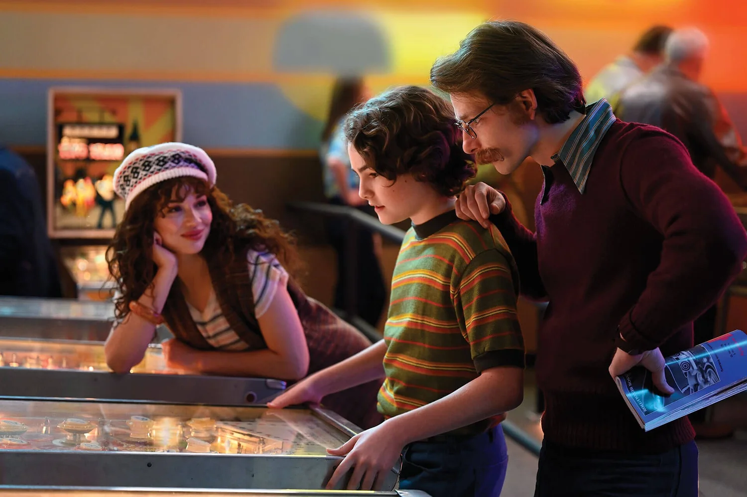 Three people in 1970s clothes gather at a pinball machine in a warmly lit arcade: A smiling young woman in a knit cap, a boy in a striped shirt at the flippers, and a mustached man in glasses.